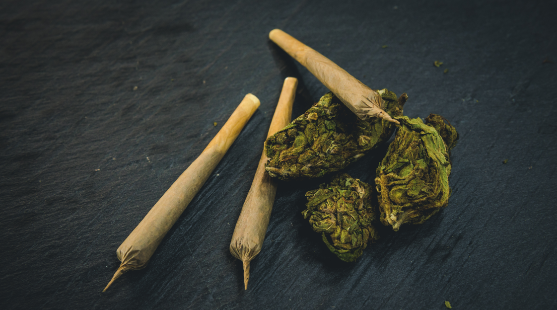 Three rolled joints and several cannabis buds on a dark surface.