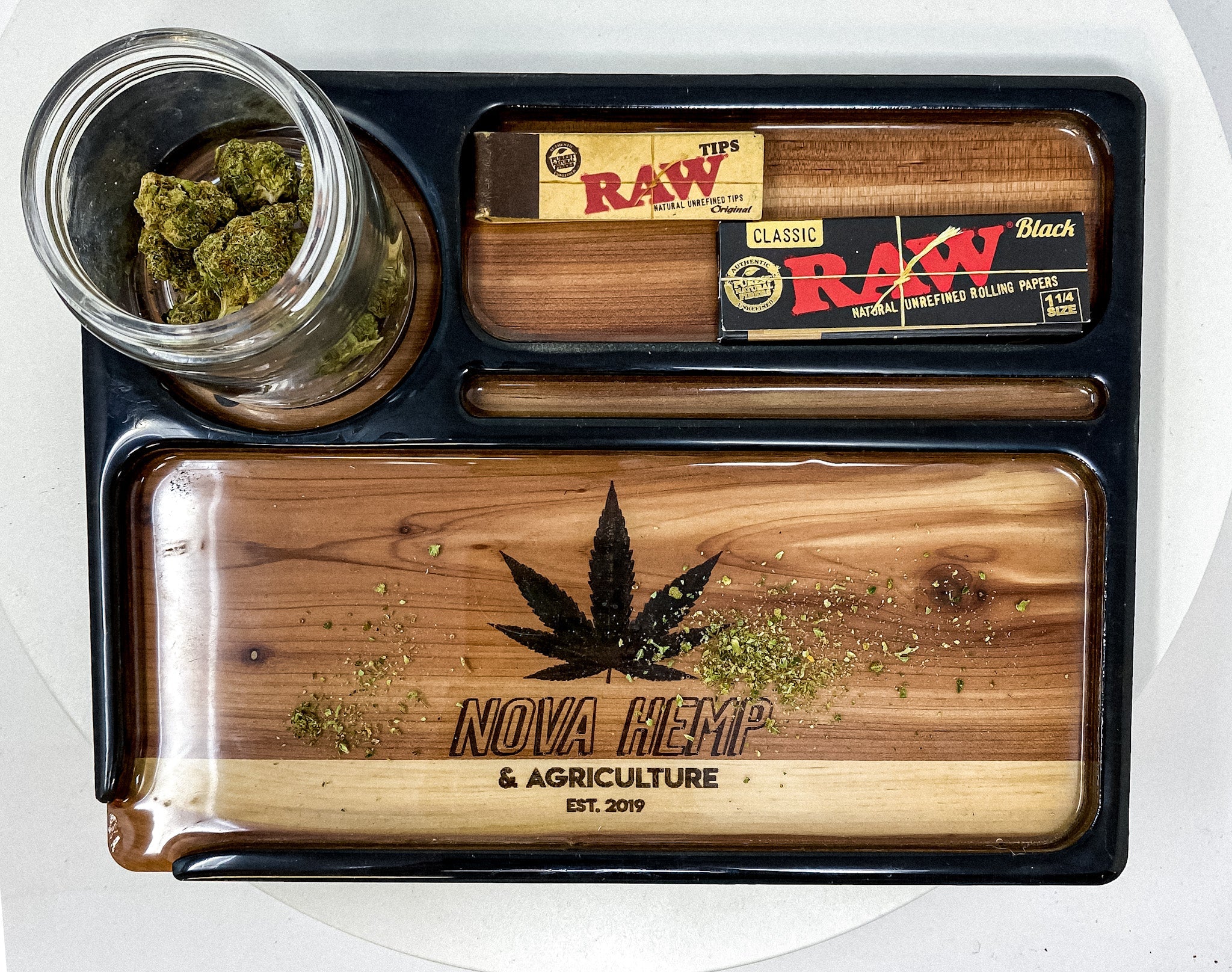 A rolling tray with a jar of cannabis buds, RAW rolling papers, and a wooden design of NOVA HEMP logo. 