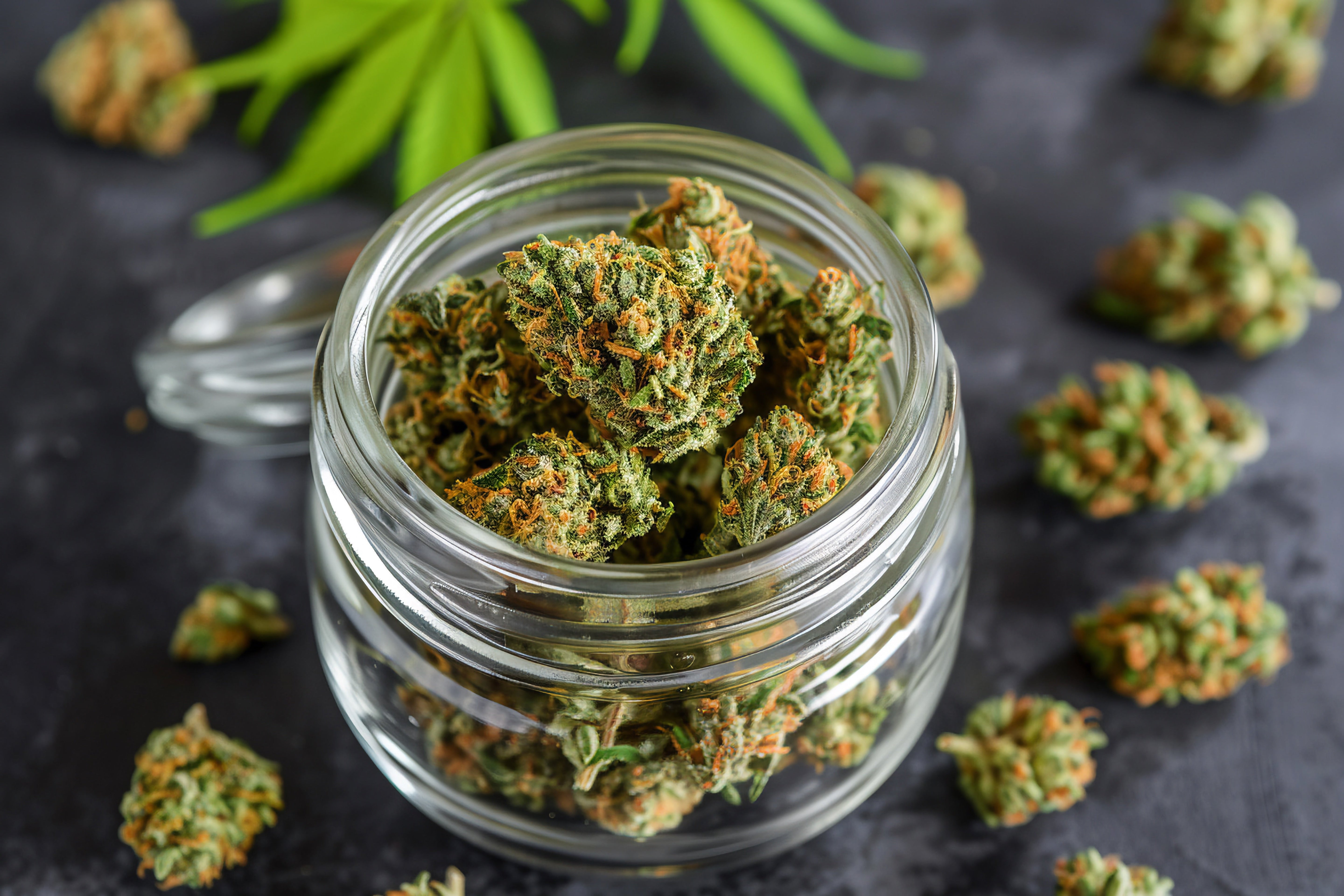 A glass jar filled with green cannabis buds, with a few buds scattered around. 