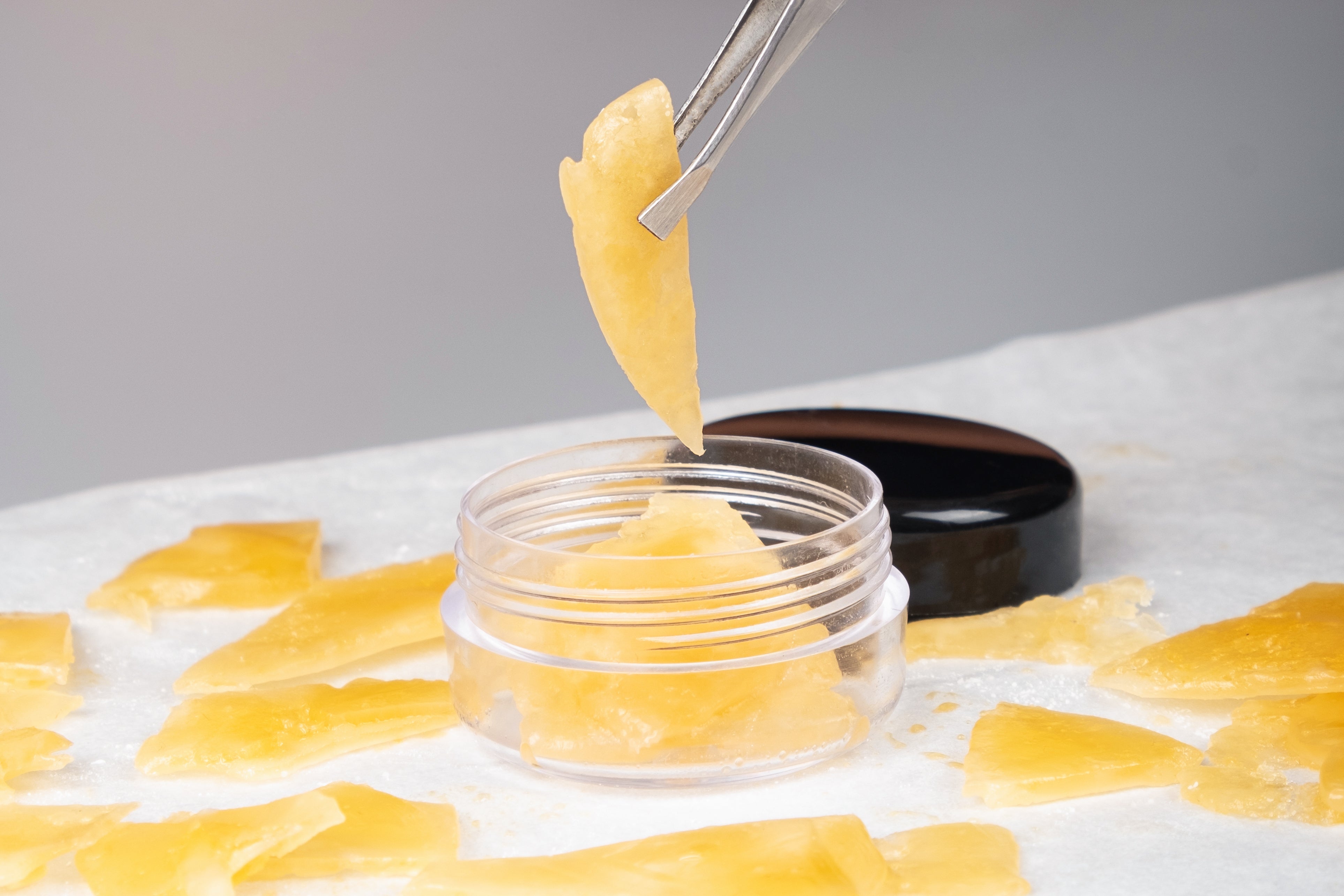 A tweezers holding a translucent, golden shard above a small clear jar filled with similar shards. 