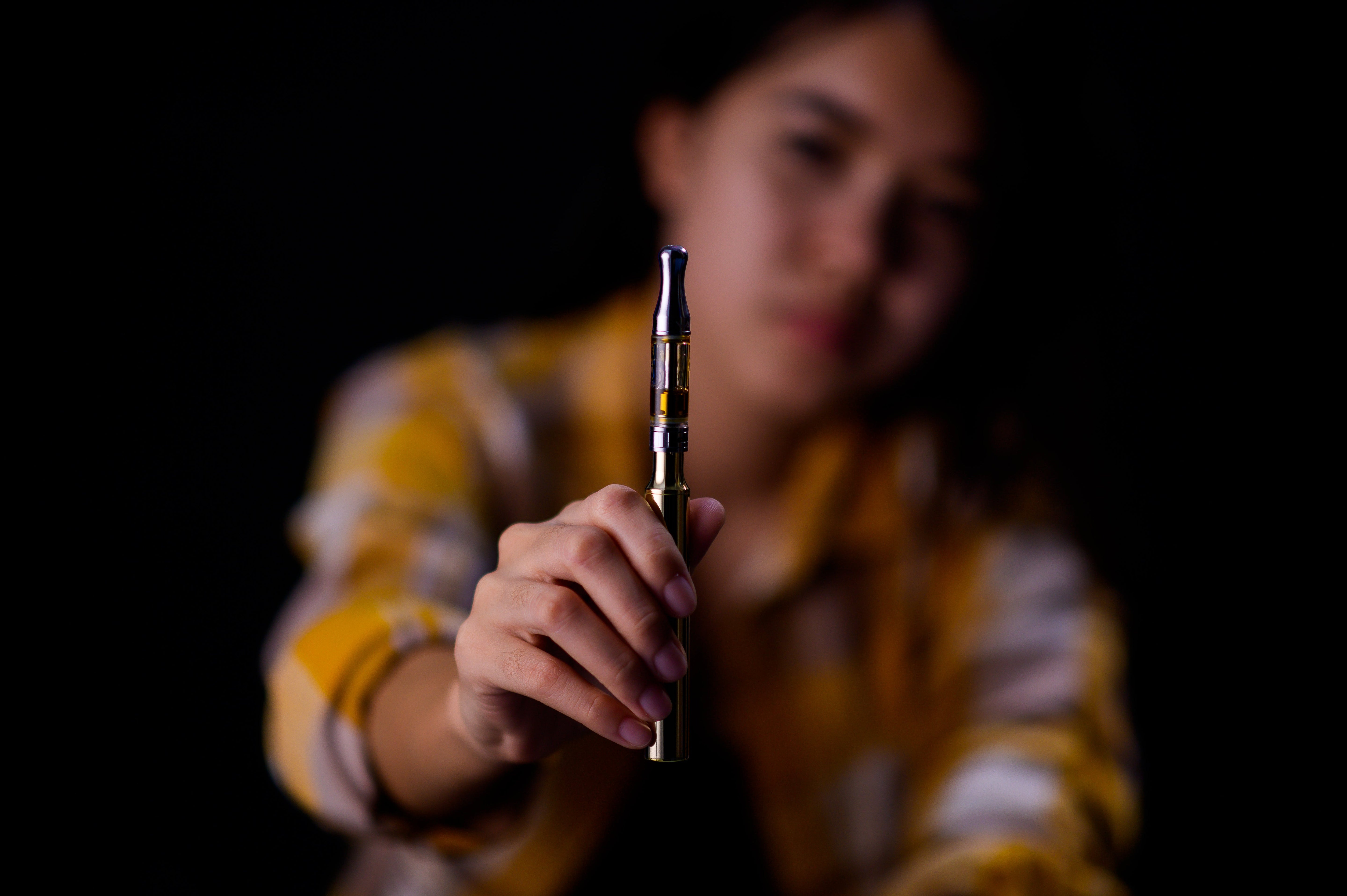 A person holding a gold vape pen with a clear tank, blurred background. 
